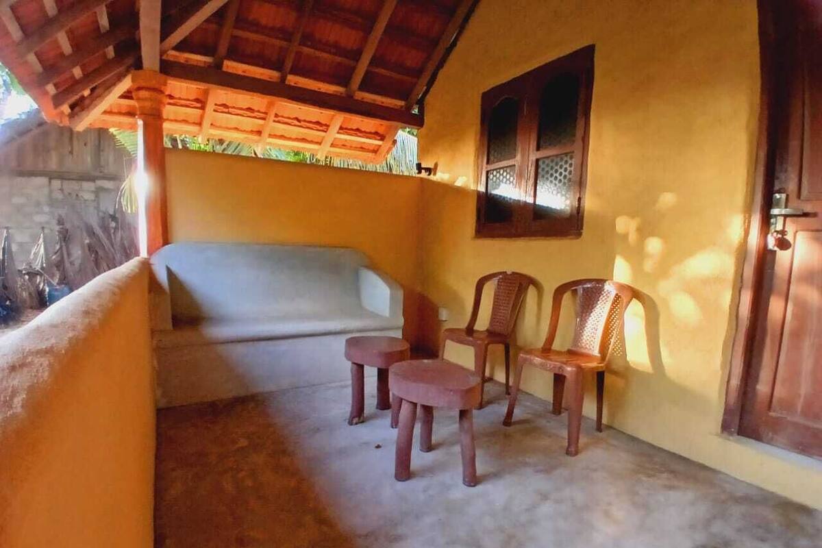 A room with a concrete floor, a bed with a wooden headboard, two small stools, and two wooden chairs.