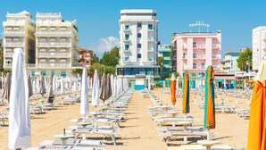 Μια παραλία με σειρές από ξαπλώστρες και ομπρέλες, κτίρια στο βάθος και έναν καθαρό, γαλάζιο ουρανό.