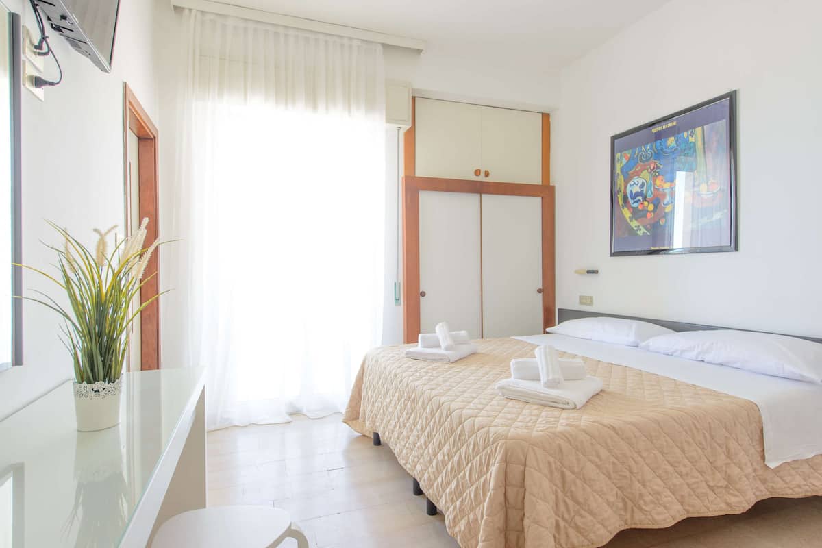 A hotel room with a bed, a painting, a plant, and a window with curtains.