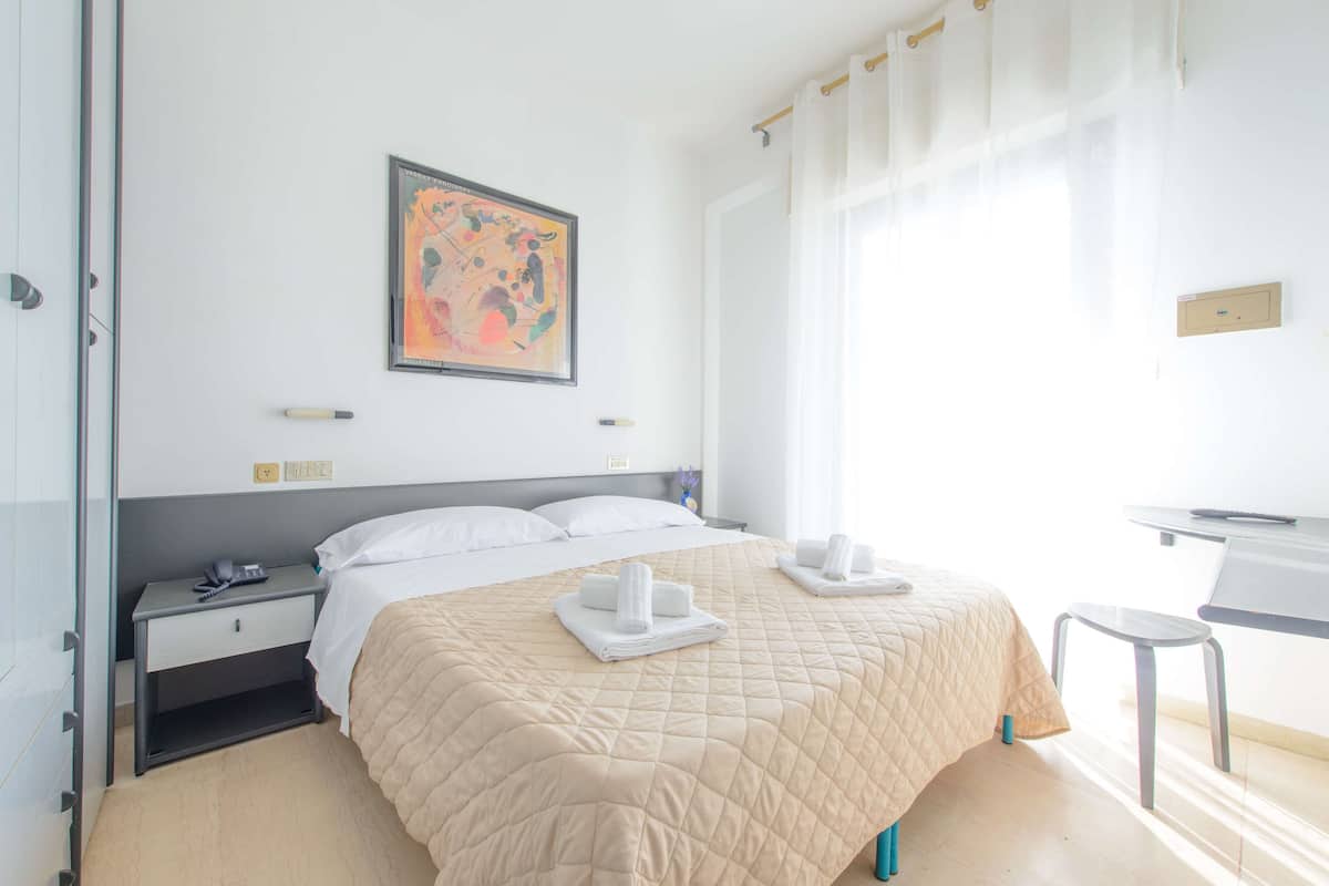 A hotel room with a large bed, a desk, and a chair. There is a painting on the wall and a window with curtains.