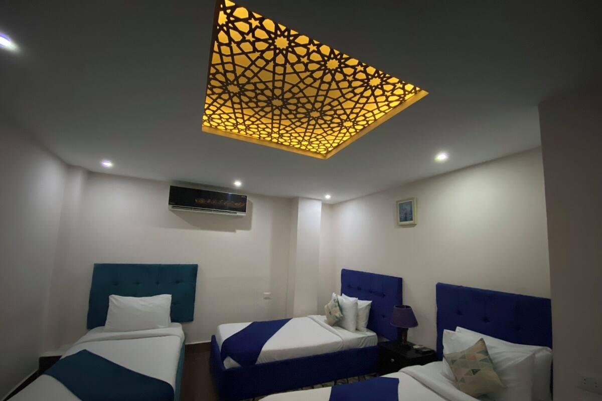 A hotel room with three beds, each with a blue headboard and white bedding. There is a decorative ceiling light fixture above the beds.