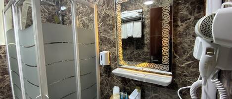 A bathroom with a glass shower enclosure, a sink with a marble countertop, a hairdryer, and a mirror with a decorative frame.