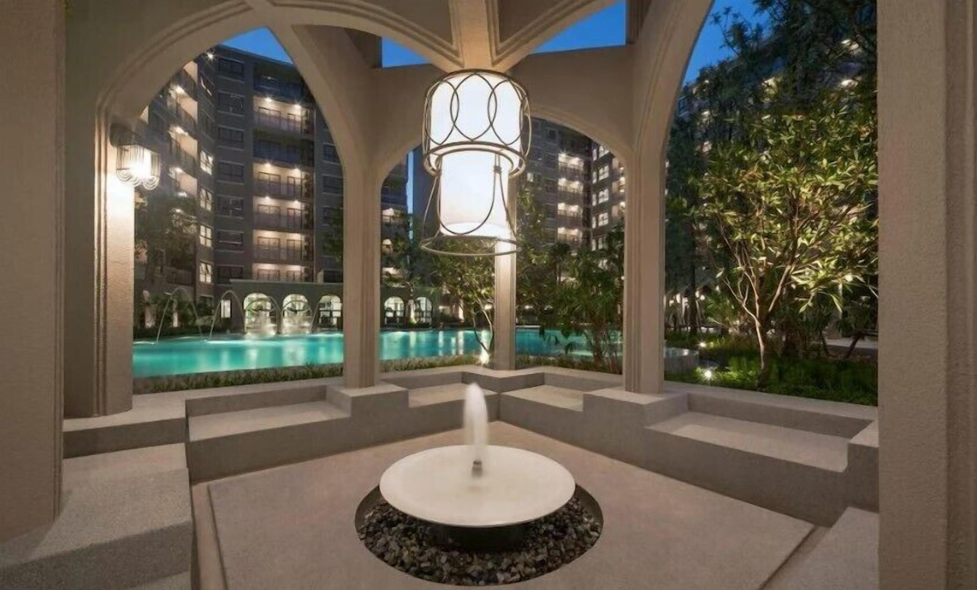 A modern outdoor pool area with a central fountain, surrounded by a covered walkway with arched ceilings.