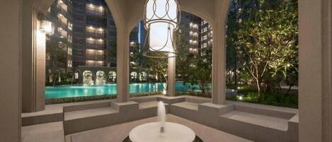 A modern outdoor pool area with a central fountain, surrounded by a covered walkway with arched ceilings.