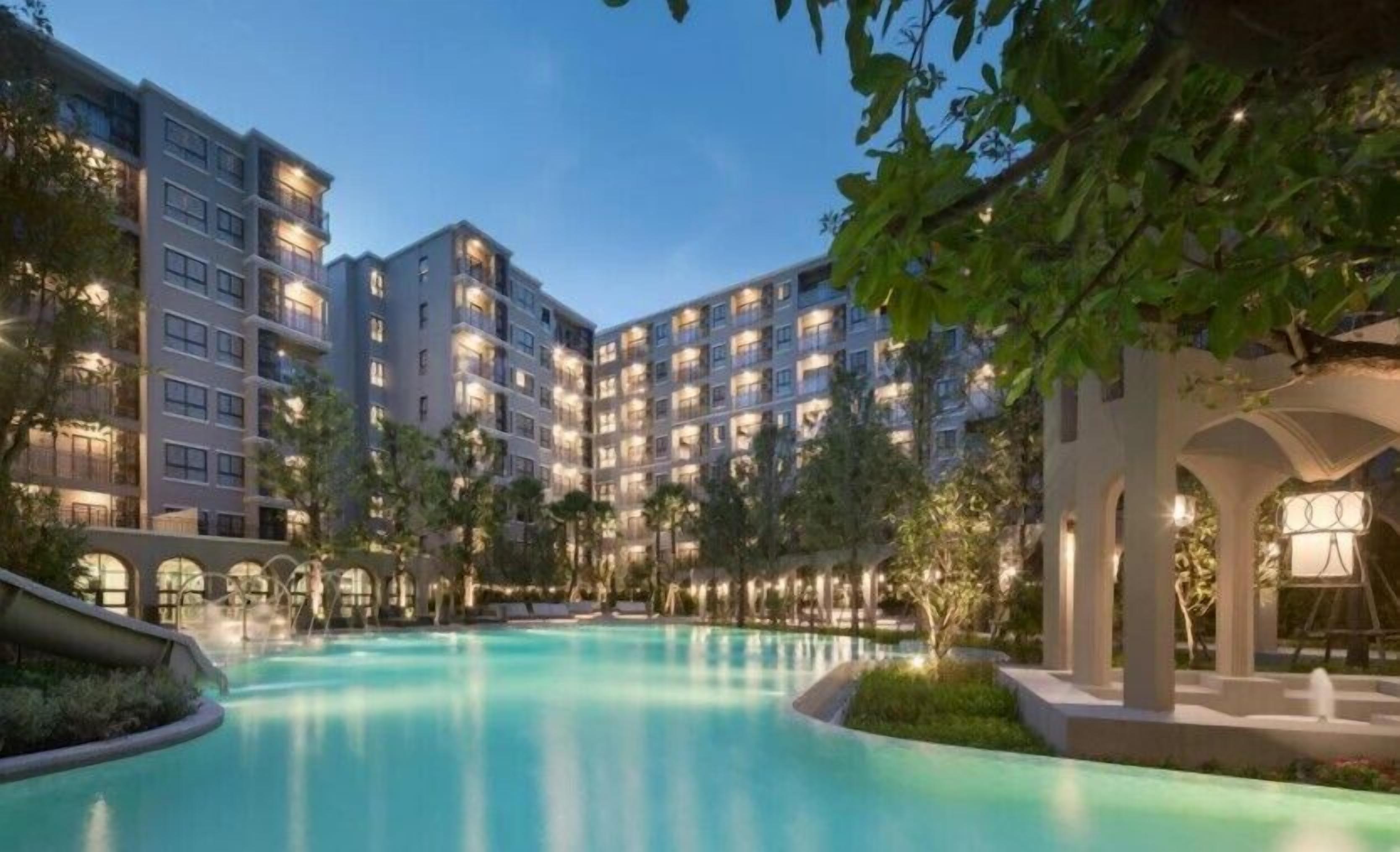 Een meerlaags hotel met een zwembad, omgeven door groen, en een overdekt gedeelte met kolommen.