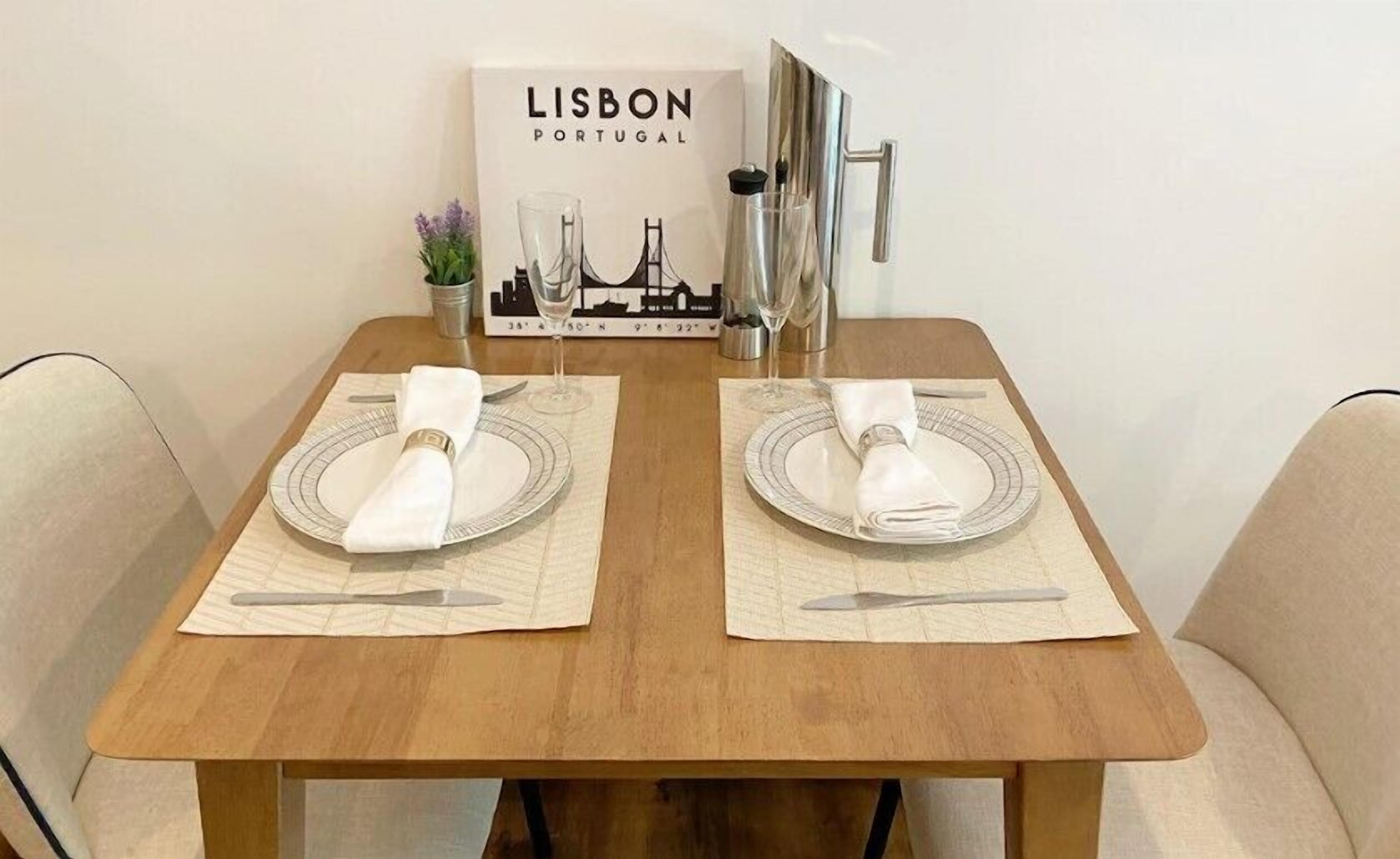 A dining table set with two plates, cutlery, and folded napkins, with a Lisbon Portugal sign in the background.
