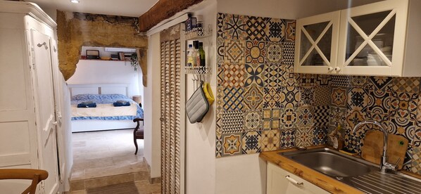 A narrow hallway leading to a bedroom with a bed, a kitchen with patterned tiles, and a wooden door.