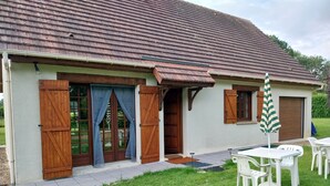 A building with a tiled roof, wooden shutters, and a patio area with white plastic chairs and a striped umbrella. - Very quiet country house (Les Monts du Roumois)