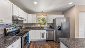 Una cocina moderna con armarios blancos, electrodomésticos de acero inoxidable, encimeras de granito y una ventana con vistas a árboles.