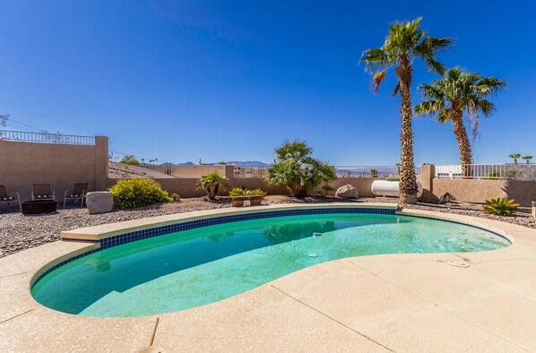 Ein Pool mit geschwungener Kante, umgeben von einer sandfarbenen Terrasse, Palmen und blauem Himmel.