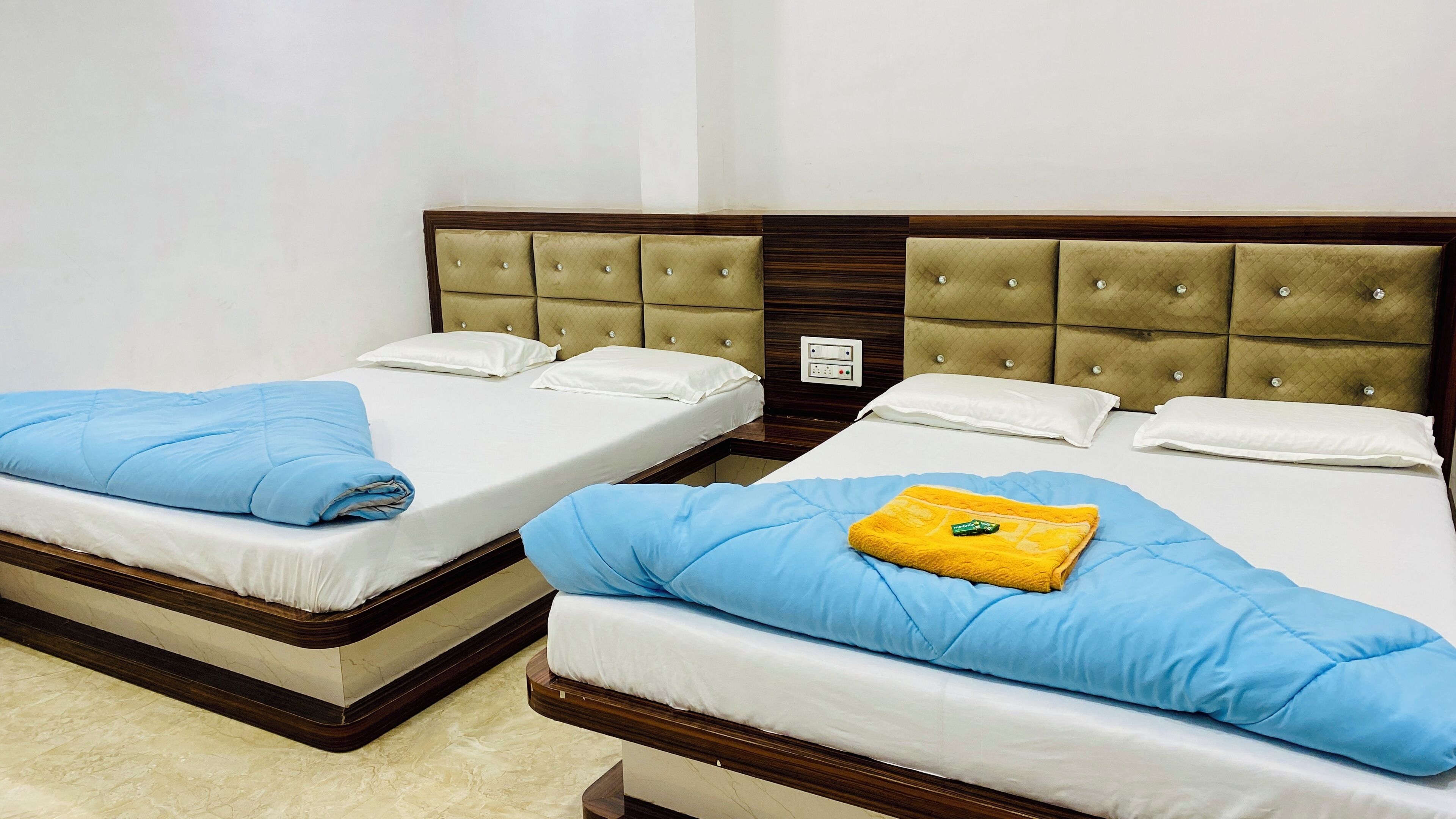 Duas camas de casal com cabeceiras de madeira e roupa de cama branca, uma pequena mesa de cabeceira e uma tomada na parede.