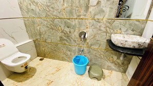 A bathroom with a marble wall, a toilet, a sink with a decorative basin, and a blue bucket. - Hotel Radhe Shree (Khandwa)