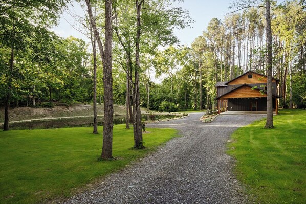 A wooden cabin with a gravel driveway, surrounded by a forested area.