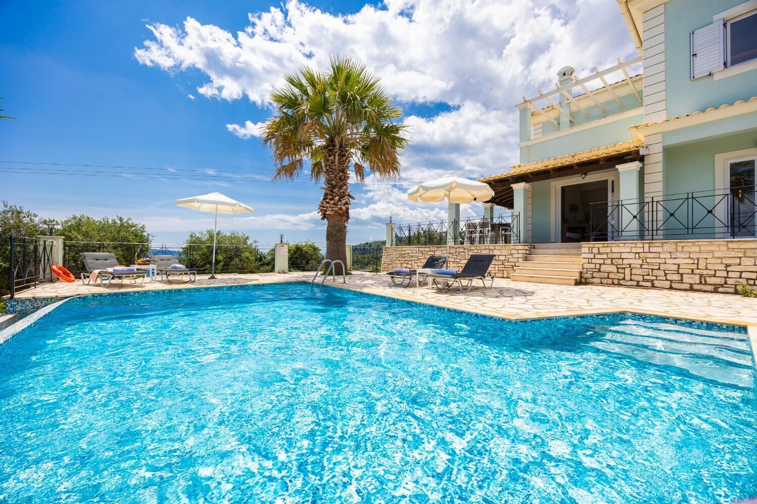 A swimming pool with lounge chairs and umbrellas, surrounded by a stone wall and a building with a balcony.