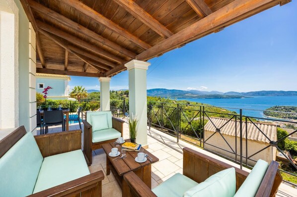 A covered outdoor seating area with wooden furniture and a view of the sea. - Villa Eleni Agios Stephanos (Άγιος Στέφανος)