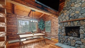 Ein Blockhaus-Interieur mit Kamin aus Naturstein, Esstisch aus Holz, Bänken und einem großen Fenster mit Jalousien.