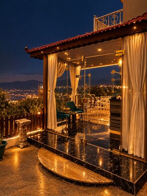 A terrace with a view of a cityscape at night, featuring a glass balustrade, a wooden deck, and a seating area with a table and chairs. - Eighth Wonders Hotel (Wadi Musa)