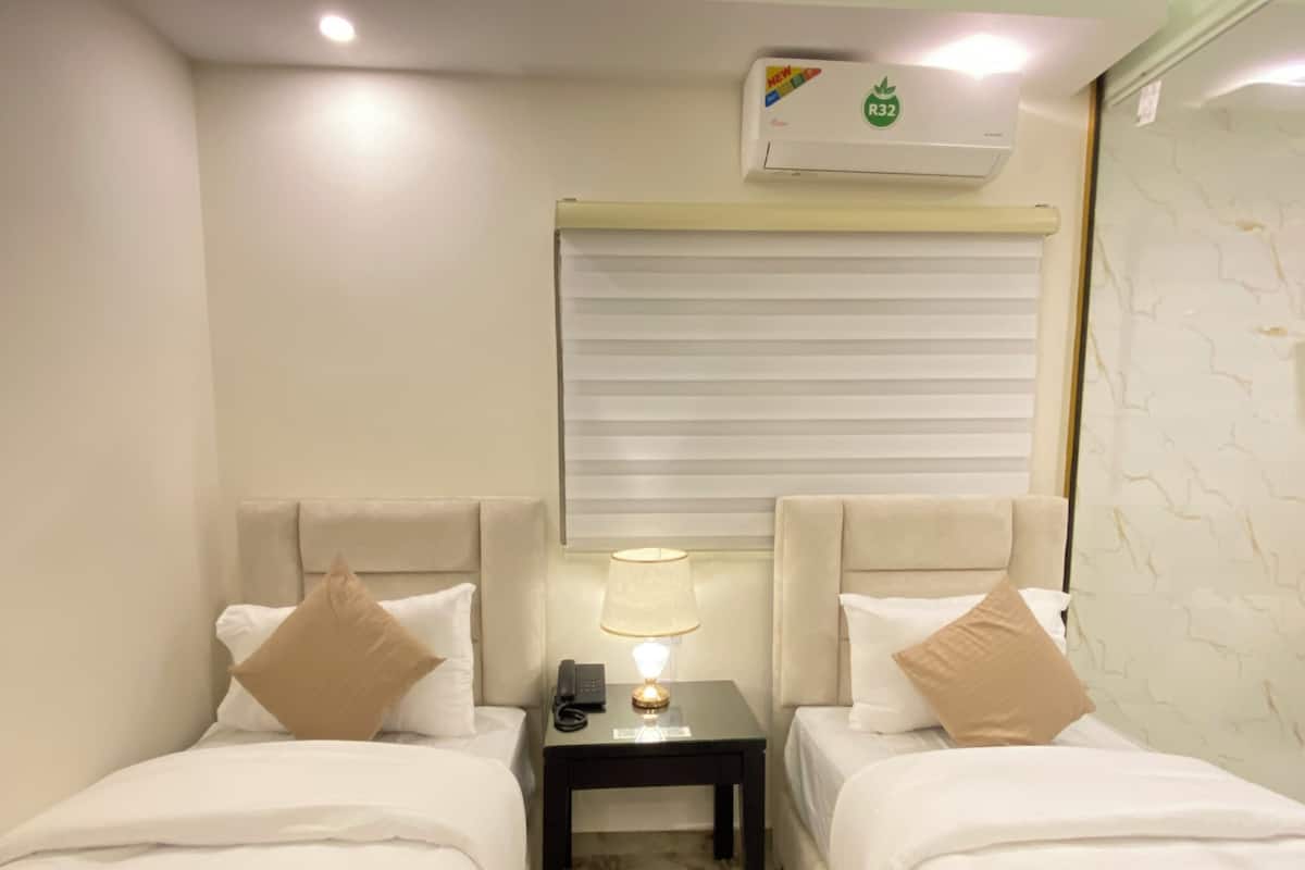 A hotel room with two beds, a nightstand with a lamp, a phone, and a window with blinds.