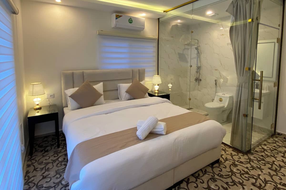 A hotel room with a large bed, bedside lamps, a glass-enclosed shower, and a marble-patterned floor.