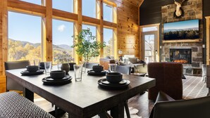 A wooden interior with a dining table set for four, a fireplace, and a large window overlooking a forested landscape. - Stunning View | Game Room Fun | Hot Tub & Fire Pit (Sevierville)