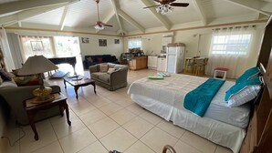 Une pièce spacieuse avec un plafond haut, un lit recouvert d’une couverture bleue, un canapé, un fauteuil, une petite table avec une lampe et un coin cuisine.