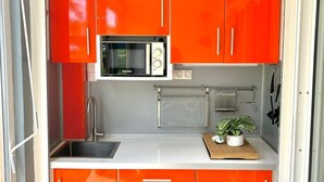 Een compacte keuken met helder oranje kastjes, een magnetron, een spoelbak en een klein aanrecht met een potplant.