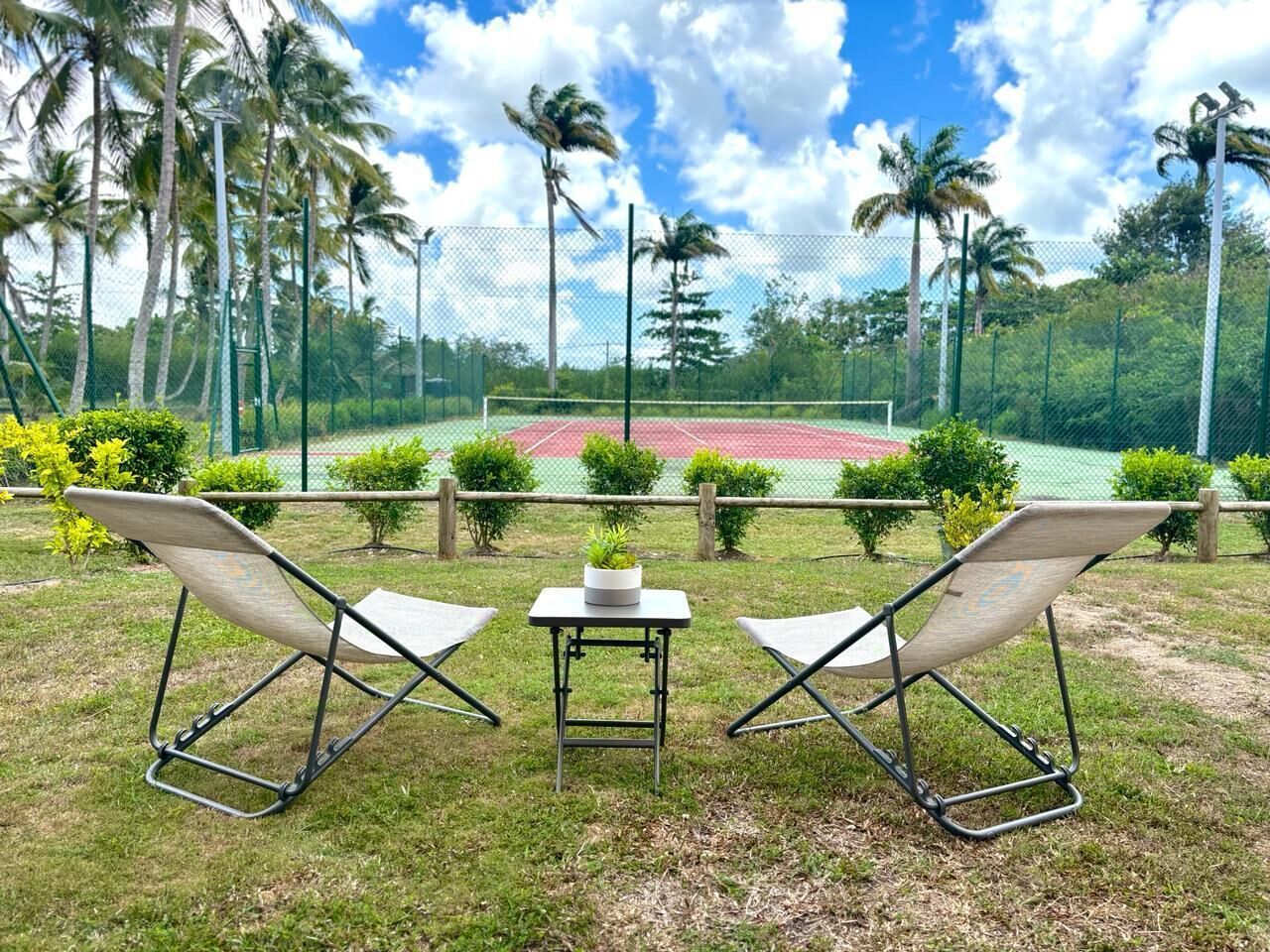 Dos sillones de descanso y una mesita con una planta en maceta en una zona con césped, con una cancha de tenis al fondo.