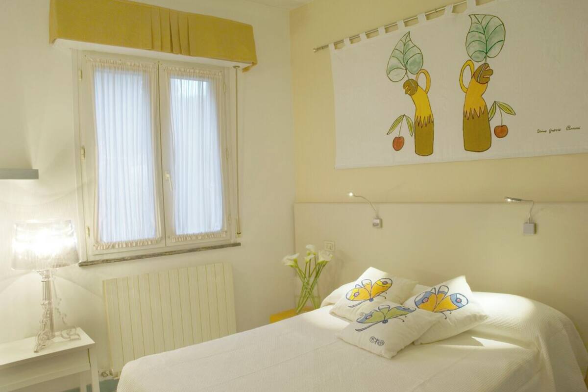 A bedroom with a bed, a window with curtains, a bedside table with a lamp, and a wall with a framed picture.