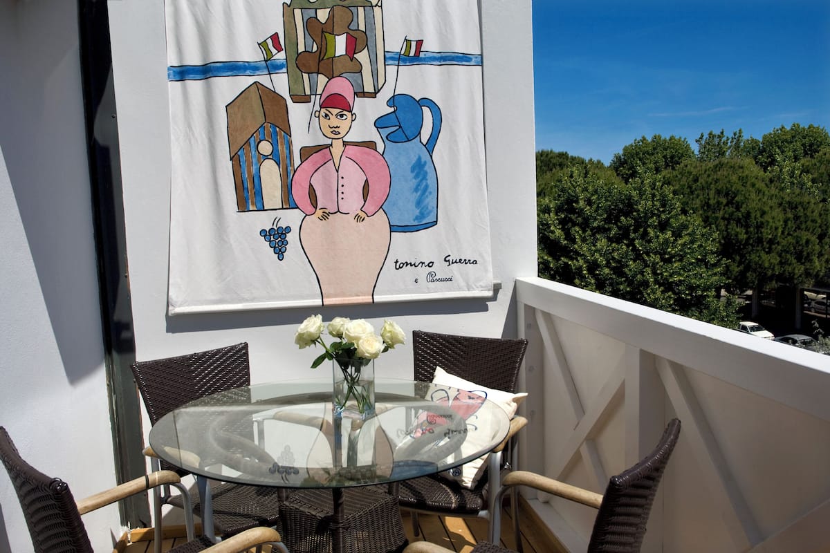 A balcony with wicker chairs, a glass table, a vase with flowers, and a wall mural.