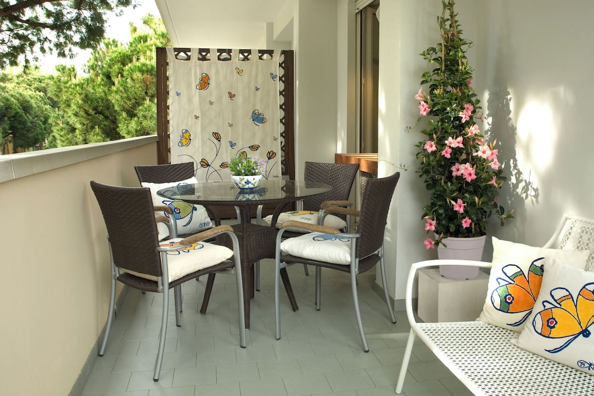 A balcony with a round table, wicker chairs, a potted plant, and a white sofa with butterfly pillows.
