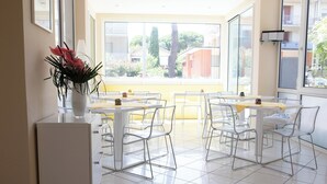 A bright, sunlit dining area with white tables and chairs, a vase of flowers on a white cabinet, and a view of a residential area through large windows. - Hotel Eros Residence (Cervia)