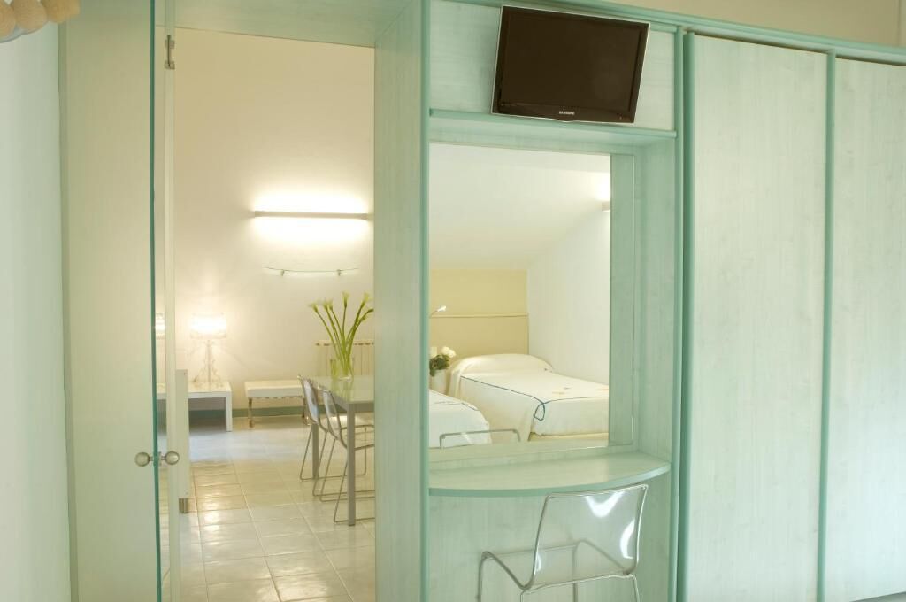 Habitación de hotel con separador de vidrio, cama, mesita de noche, silla y televisor montado en la pared.