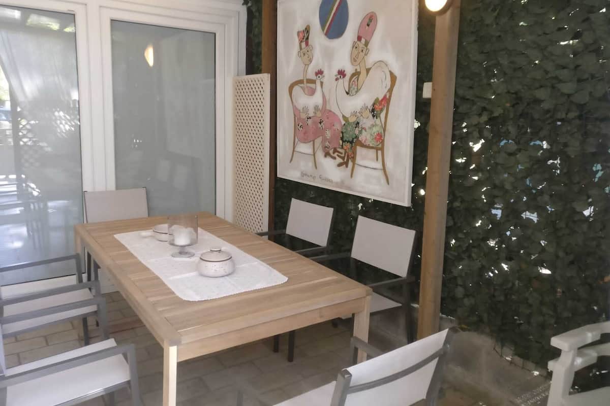 A dining area with a wooden table, white chairs, and a glass partition. There is a painting on the wall and a green leafy wall behind the table.