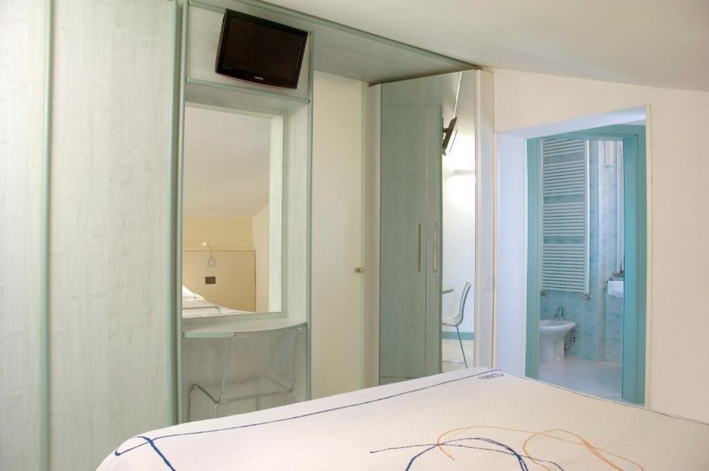 A bedroom with a bed, a TV mounted on a glass partition, a bathroom with a toilet and sink, and a window with blinds.