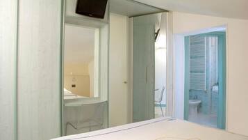 A bedroom with a bed, a TV mounted on a glass partition, a bathroom with a toilet and sink, and a window with blinds.