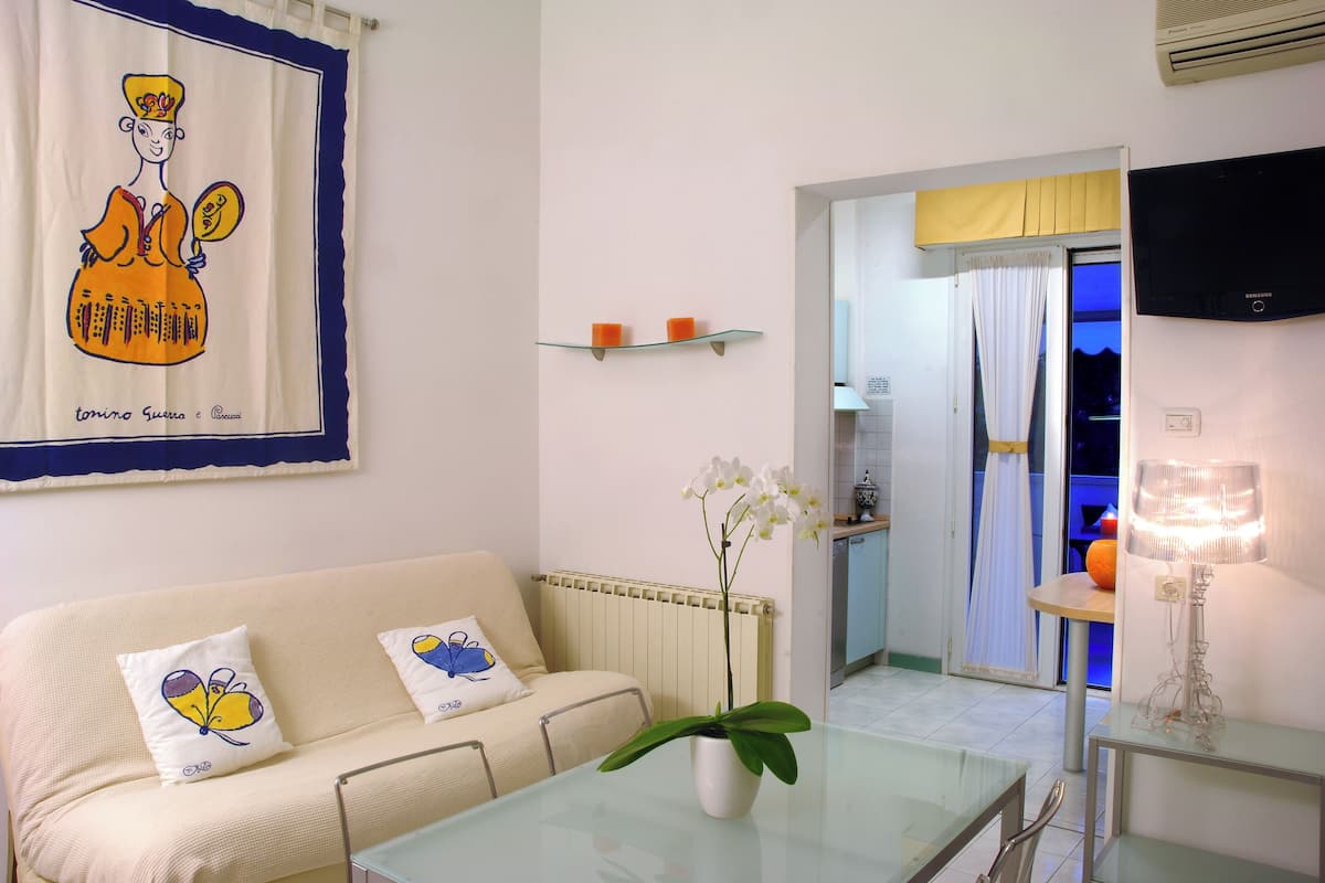 A living room with a sofa, a glass table, a potted orchid, and a TV mounted on the wall.
