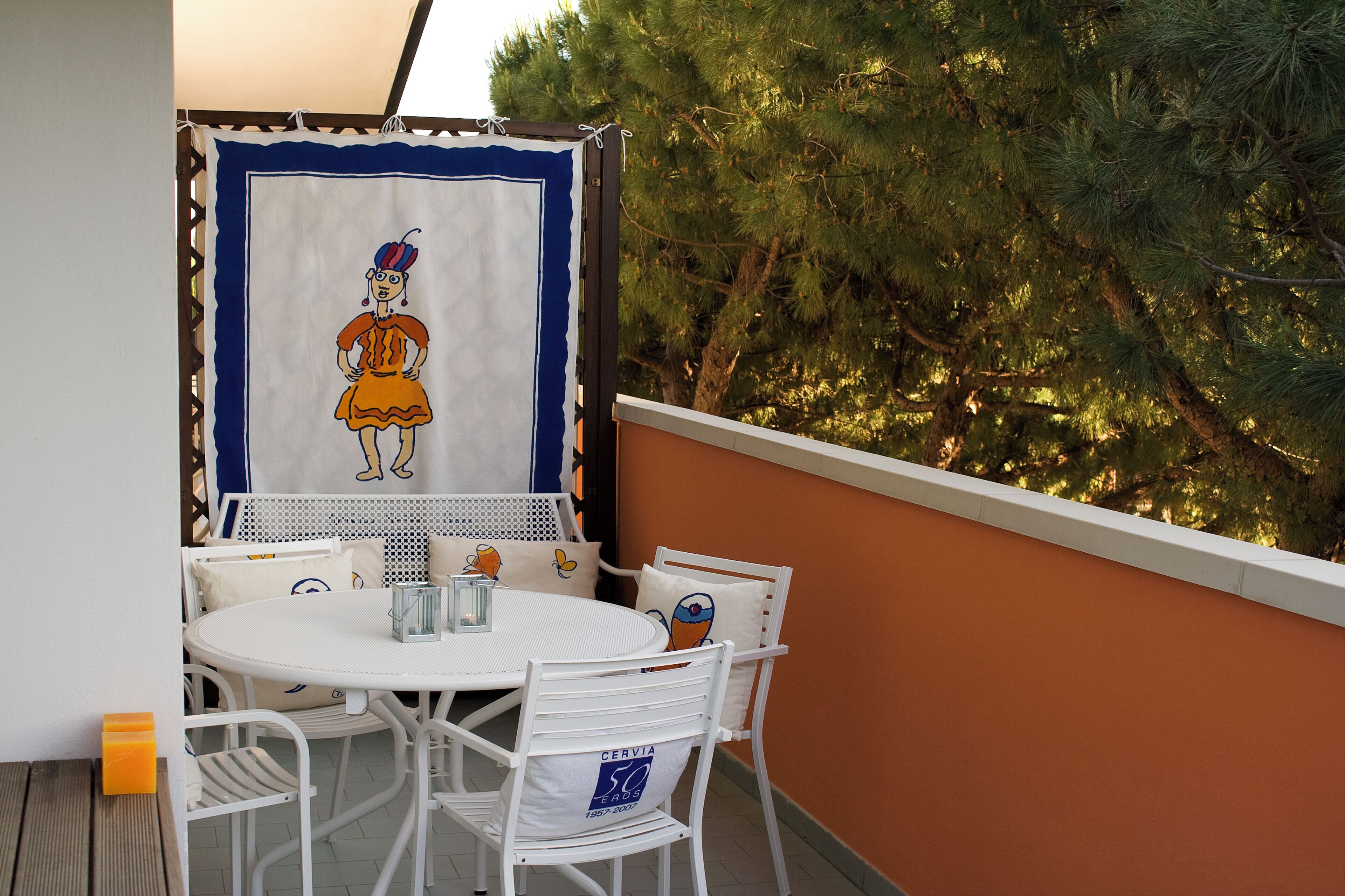 A balcony with a white table and chairs, a decorative cloth with a cartoon character, and a view of trees.