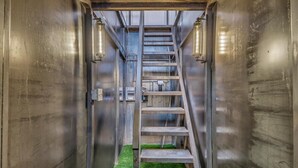 Un couloir étroit et métallique, avec un escalier en métal et, au bas, un tapis d’herbe artificielle verte.