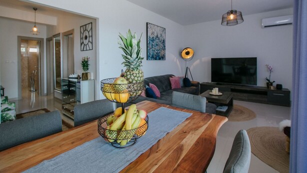 A modern living room with a wooden dining table, grey sofas, a TV, and a fruit bowl. - Scenic Sea View Apartment Trogir - Two Bedroom Apartment with Balcony and Sea View (Trogir)