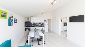 A modern kitchen with white cabinets, a black countertop, and a microwave. There is a dining area with a white table and chairs, and a TV mounted on the wall. - Two Bedroom Apartment with Balcony (Kastel Novi)
