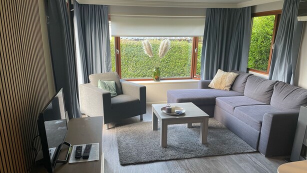 Una sala de estar moderna con sofá, sillón y mesa de centro. Hay un televisor sobre un mueble bajo y una ventana con cortinas.