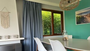 Un comedor con una mesa, sillas, un tapiz de macramé, una ventana con cortinas y una lámpara colgante.