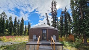 A yurt with a wooden deck and two chairs, surrounded by a forest. - Premium Comfort in the Wilderness (Nabesna)
