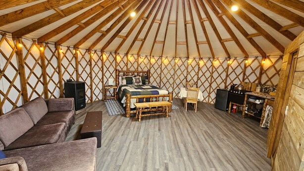 A cozy yurt interior with a central bed, wooden furniture, and a dining area. - Premium Comfort in the Wilderness (Nabesna)