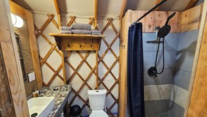 A compact bathroom with a white toilet, a sink with a patterned countertop, a shower area, and a wooden shelf above the toilet. - Premium Comfort in the Wilderness (Nabesna)