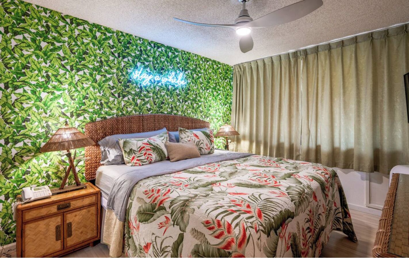 Une chambre à coucher comprenant un lit, une table de chevet, une lampe et un mur orné d’un motif végétal.