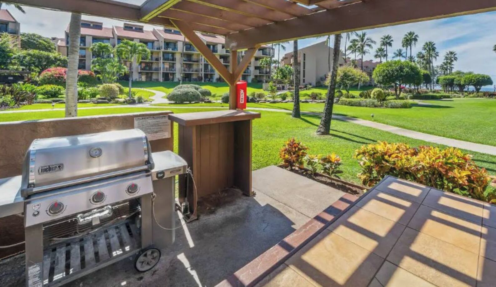 An outdoor patio with a built-in grill, a fire extinguisher, and a view of a well-maintained lawn and buildings in the background.