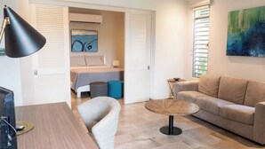 Una sala de estar moderna con un sofá, una mesa de centro redonda y un televisor. Se ve una cama al fondo, con un cuadro en la pared.
