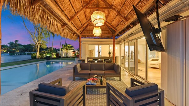 An outdoor lounge area with a pool, a sofa, and a TV screen. - 702A | Waterfront | Sleeps 8 | Heated Pool |Tiki Hut |Fire Pit |by DelrayNightly (Delray Beach)