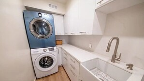 Un espace réduit dédié à la buanderie, comprenant un lave-linge bleu et blanc, un évier avec un robinet et un petit panier.